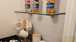 A kitchen counter with Torani syrup bottles, a coffee jar, sugar and tea containers, a coffee machine, and a tray of donuts.