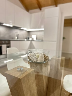 Une cuisine moderne avec des meubles blancs, une table à manger en verre agrémentée d’un centre de table en verre transparent, et un élément décoratif en bois sur la table.