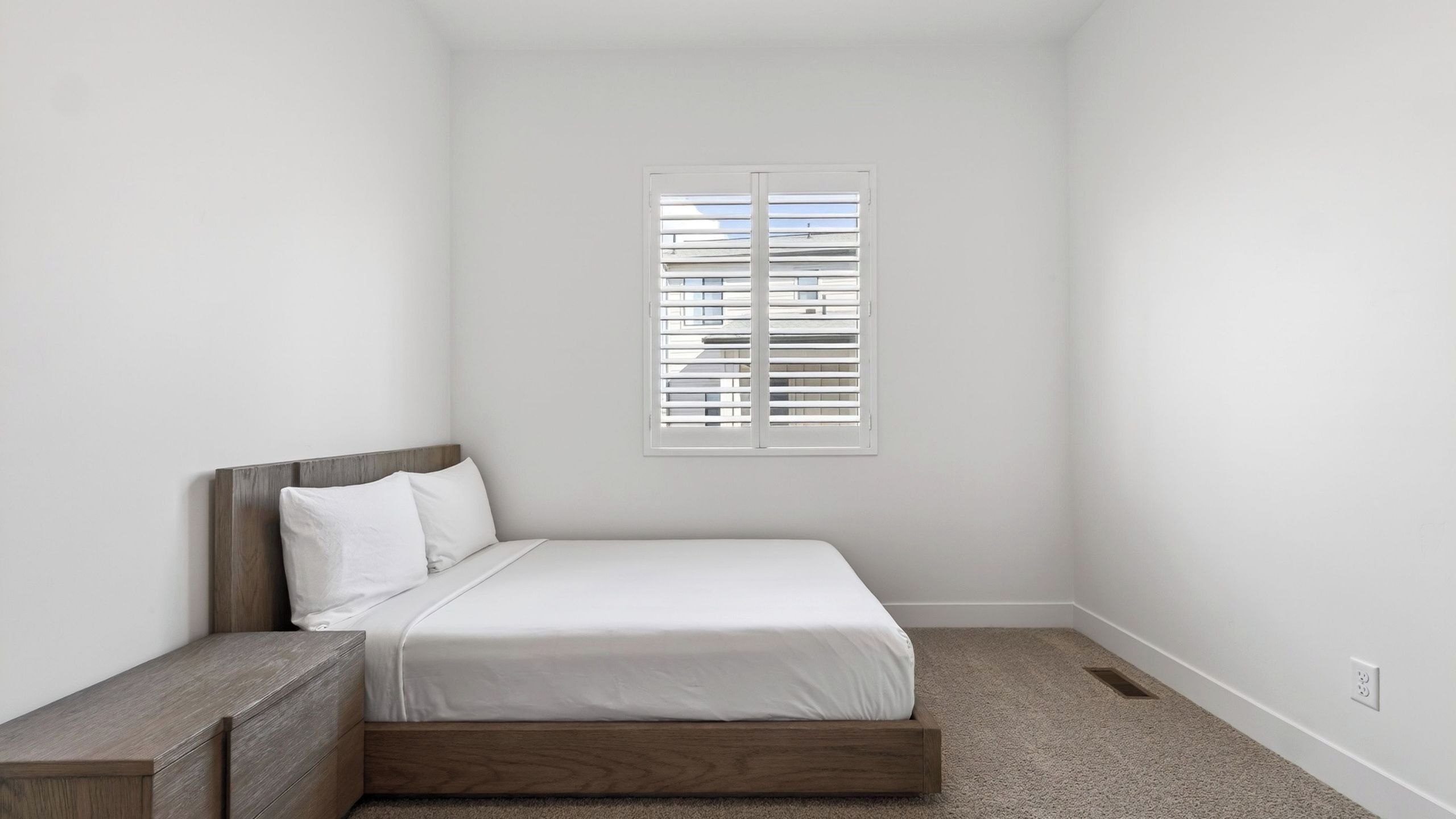 A bedroom with a single bed, a nightstand, a window with blinds, and a carpeted floor.