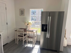 Une cuisine équipée d’un réfrigérateur en acier inoxydable, une table à manger avec des chaises blanches, une fenêtre donnant sur des immeubles et un vase rempli de fleurs posé sur la table.