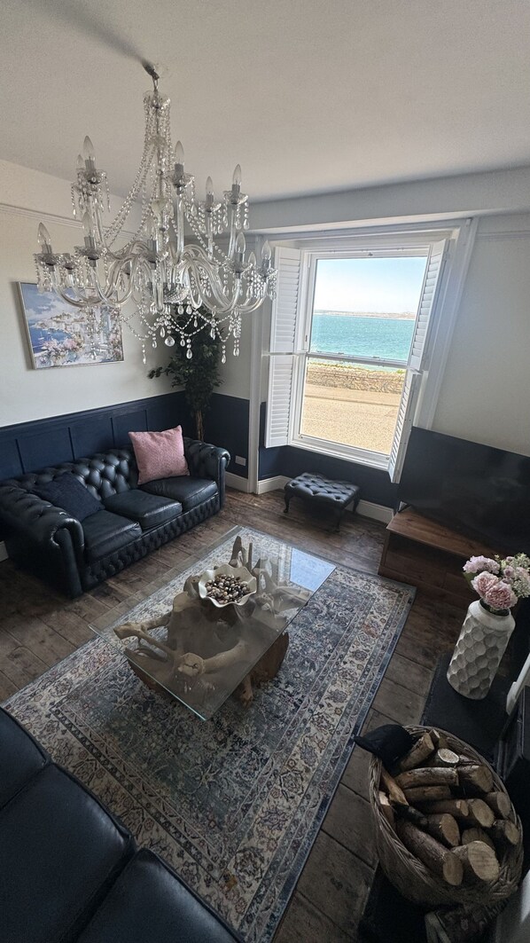 A living room with a black leather sofa, a glass coffee table, a chandelier, a painting, and a view of the sea through a window. - Minster Panorama is a beautifully restored three story Georgian Town House located on the famous "Th (St Ives)
