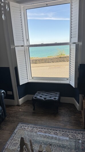 Een kamer met een erker die uitkijkt op een strand, een zwarte leren poef en een kamerbreed tapijt met dessin.