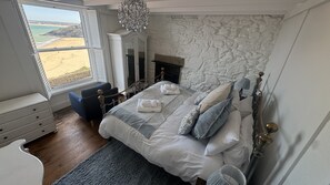Een slaapkamer met een stenen muur, een open haard, een bed met handdoeken, een raam met uitzicht op het strand en een kroonluchter.