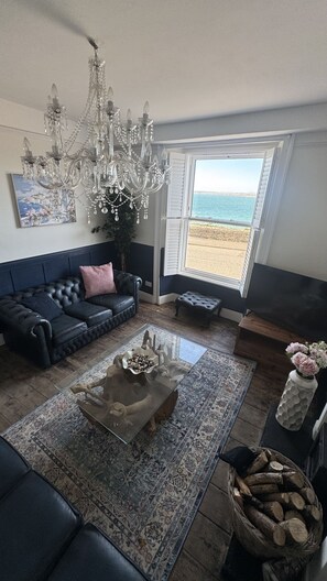 A living room with a black leather sofa, a glass coffee table, a chandelier, a painting, and a view of the sea through a window. - Minster Panorama and Minster Vista Connected is a beautifully restored three story Georgian Town Hou (St Ives)