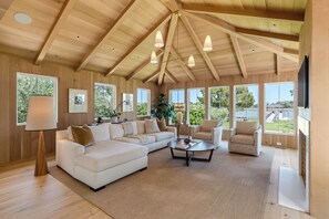 Un salon spacieux avec un grand canapé d’angle, une table basse et une cheminée. De nombreuses fenêtres laissent entrer la lumière naturelle et offrent une vue sur l’extérieur.
