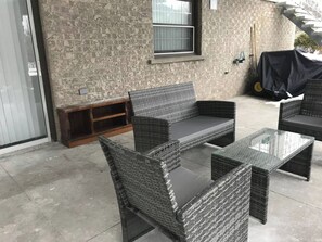 Arredamento da esterno in vimini con cuscini, un tavolo in vetro e un muro in mattoni con una finestra.