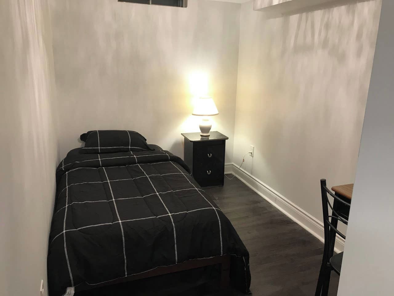 Una camera da letto piccola e poco illuminata, con un letto singolo, un comodino con una lampada e una sedia.