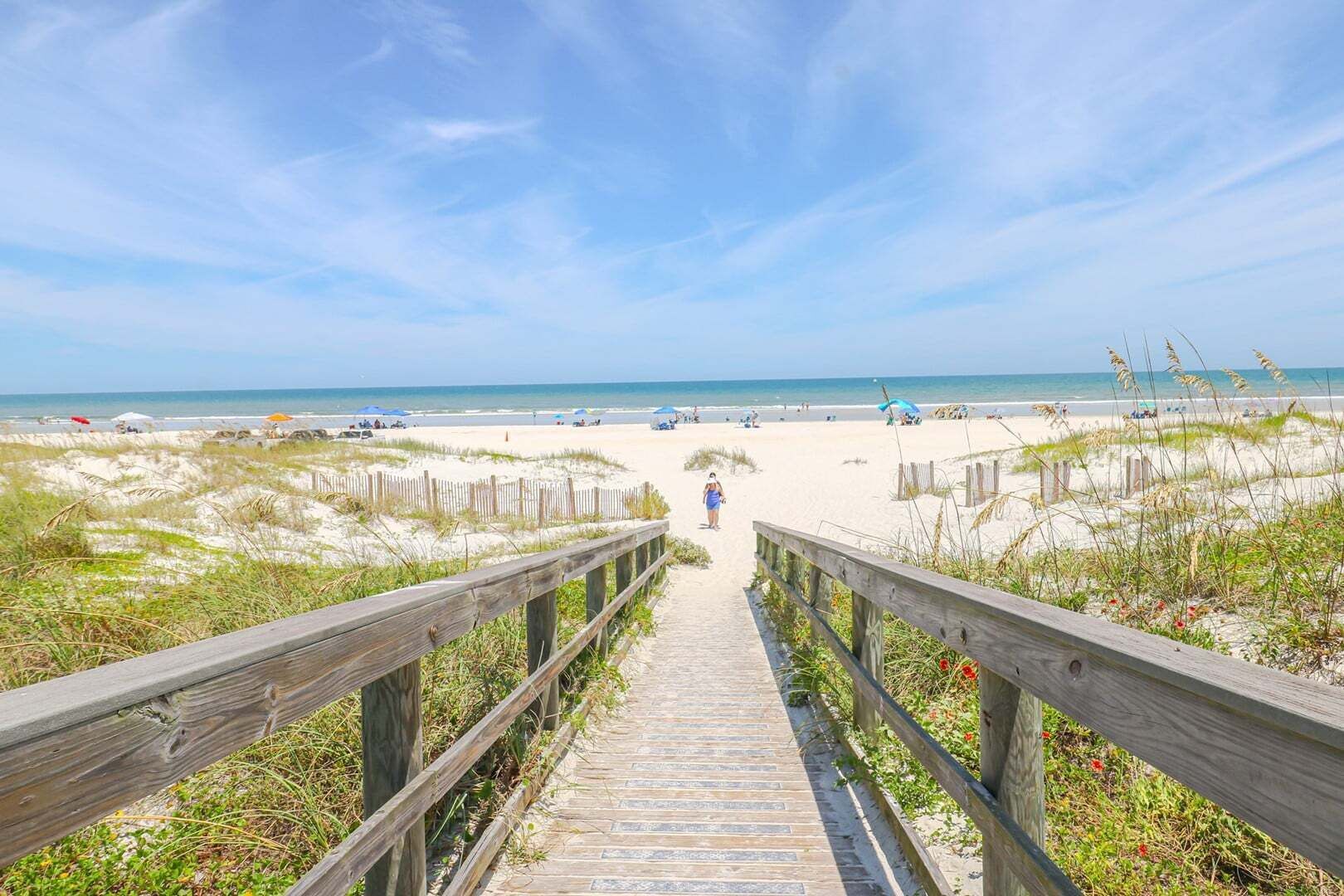 Ein hölzerner Steg führt zu einem Sandstrand mit Dünen und blauem Himmel.