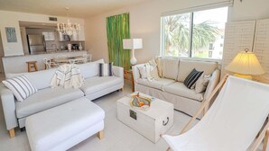 Una sala de estar moderna con un sofá, un sillón y una mesa de centro. Se observa una cocina al fondo con zona de comedor.