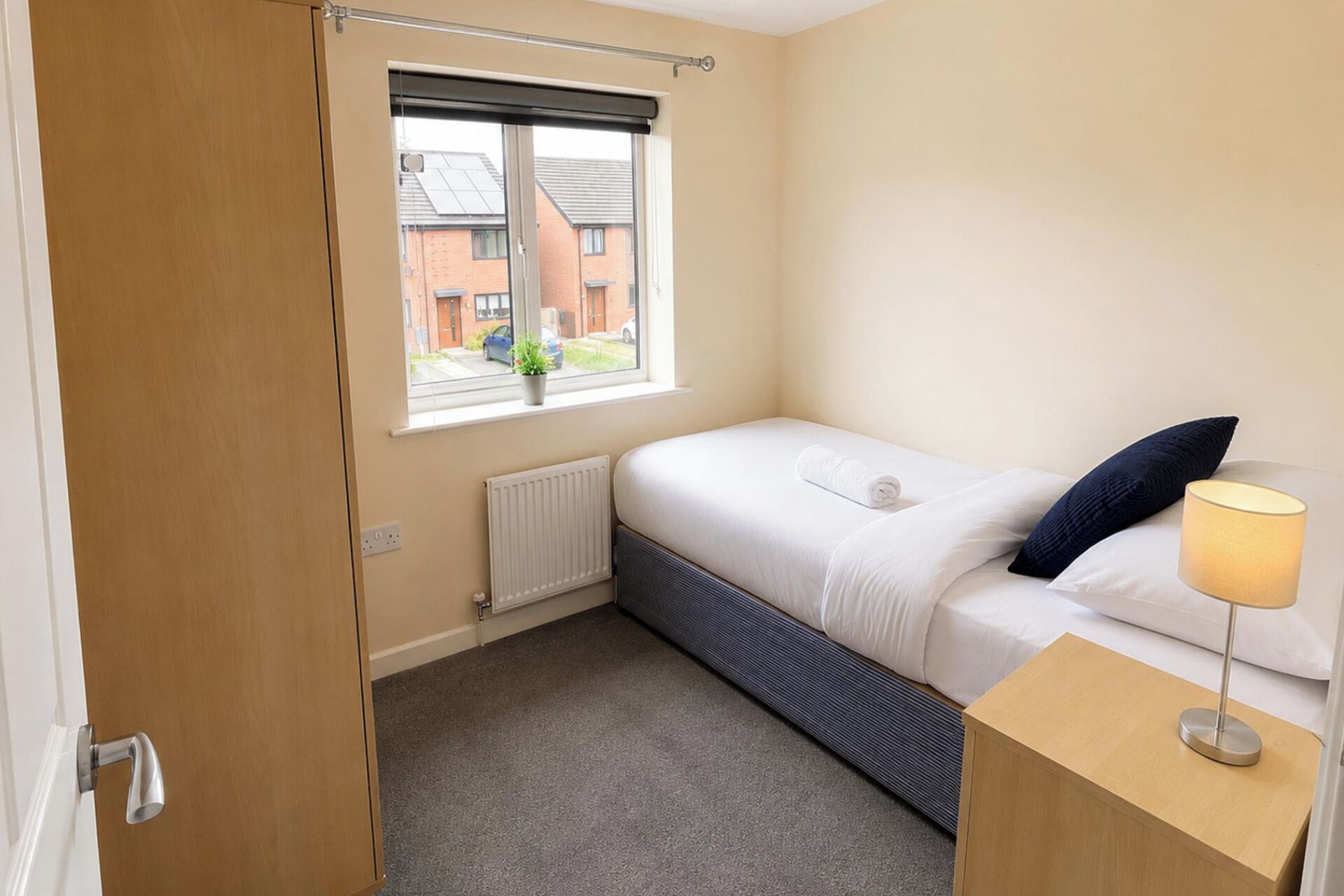 A bedroom with a bed, a bedside table, a lamp, a radiator, a window with a view of buildings, and a door.