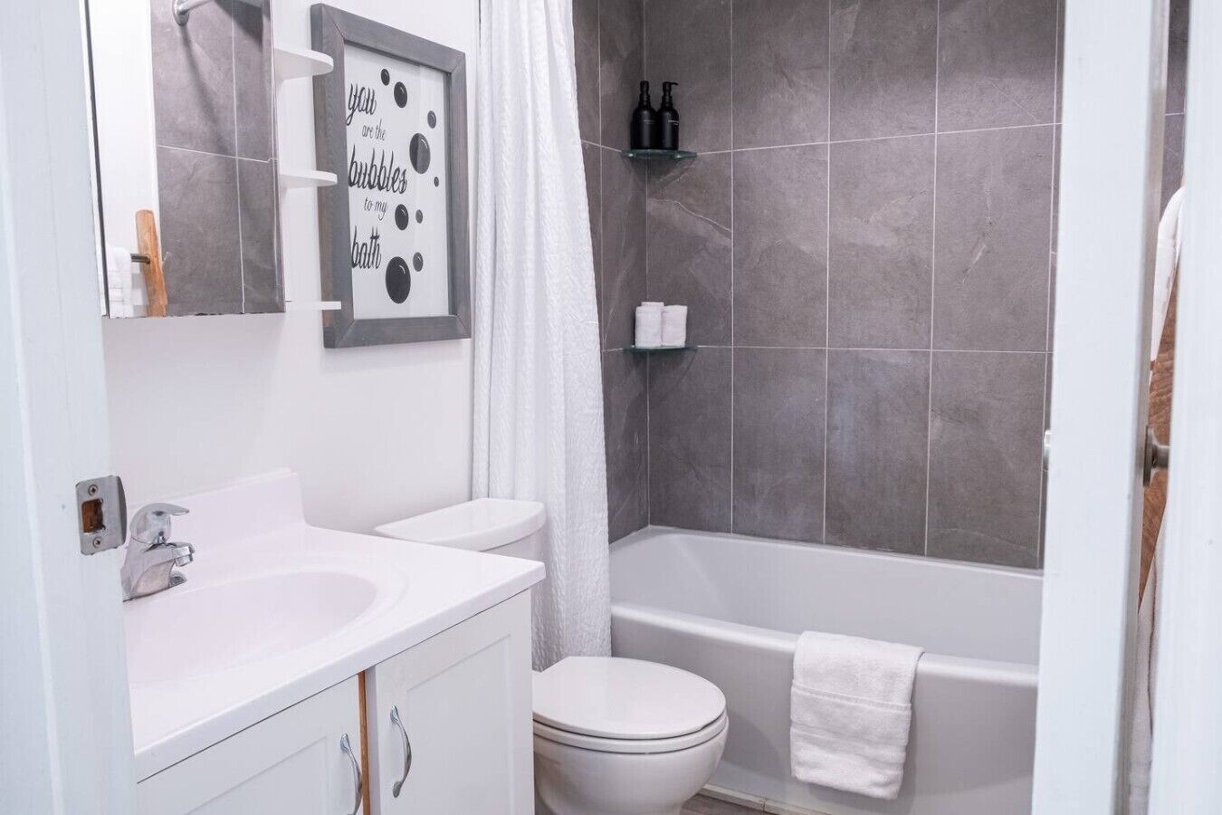 A bathroom with a white sink, toilet, and bathtub. There is a framed quote on the wall, a shower curtain, and toiletries on a shelf.