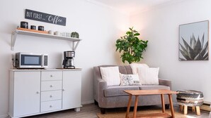 A living room with a grey sofa, a coffee machine, a microwave, a small cabinet, a plant, and a framed picture on the wall.