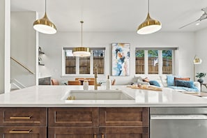 A modern kitchen with a large island, pendant lights, a dishwasher, and a dining area with a view of the backyard.