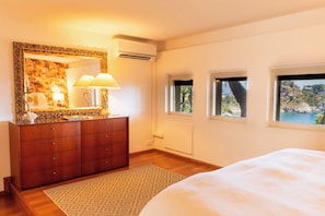 A bedroom with a wooden dresser, a mirror with a decorative frame, a bedside lamp, a bed, and three windows with a coastal view. - Villa Marina Taormina (Mazzaro')