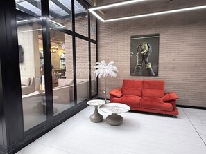Un interior moderno con un sofá rojo, dos mesas redondas, una planta decorativa blanca y un ventanal grande.