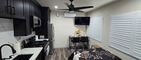 A compact kitchen with a stove, microwave, and refrigerator, a dining area with a table and chairs, a ceiling fan, and a clock on the wall.