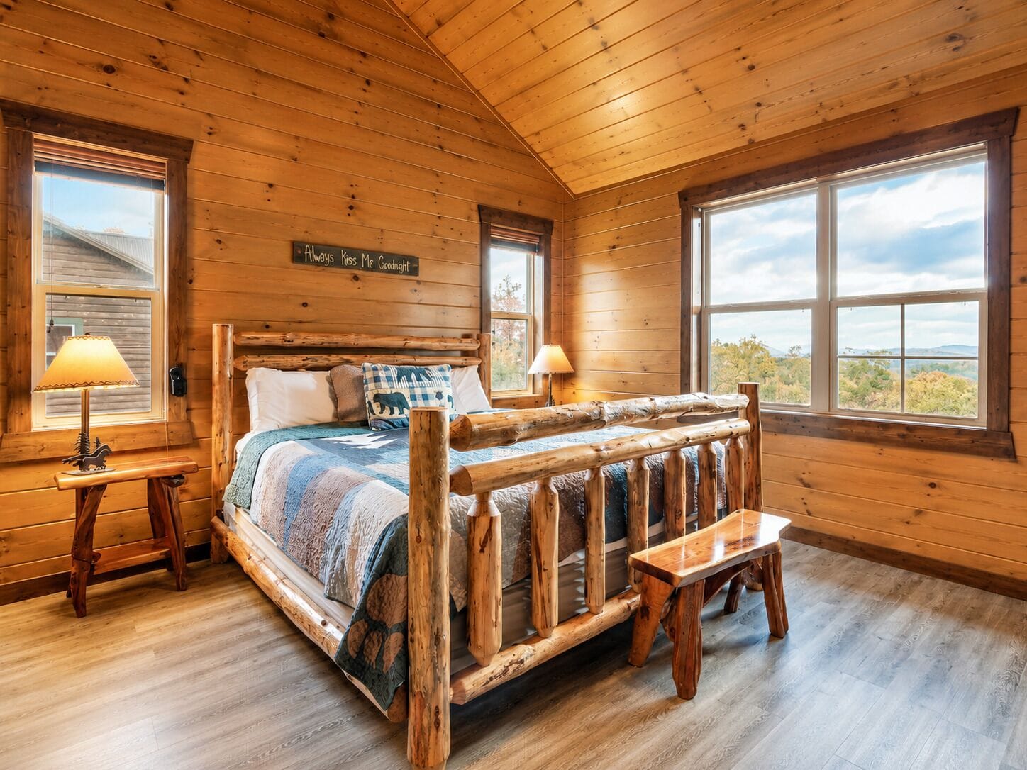 A wooden cabin bedroom with a bed, two bedside lamps, a bench, and a window with a view.