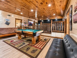 A pool table in a room with wooden walls and ceiling, a black leather couch, and a billiards-themed carpet.