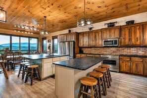 A spacious kitchen with wooden cabinets, a large island with stools, stainless steel appliances, and a view of the mountains through large windows. - Luxury Smoky Mountain Cabin w/ Indoor Pool, Theater & Panoramic Views (Sevierville)