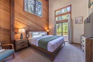 Ein Schlafzimmer mit Holz-Kopfschild, einem Bett mit weißer Bettwäsche, einem Nachttisch mit Lampe, einer Kommode und einem Fenster mit Blick auf Bäume.