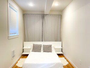 Een slaapkamer met een bed, een raam met jaloezieën, een plafondlamp, een ventilatie rooster en een wit vloerkleed.