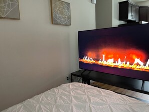 Een slaapkamer met een televisie waarop een haard te zien is, een bed met een witte dekbedovertrek en een wandmeubel.