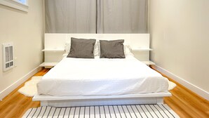 Ein Schlafzimmer mit einem Bett, zwei Nachttischen, einem weißen Kopfteil, einem Fenster mit Jalousien und einem weißen Teppich.