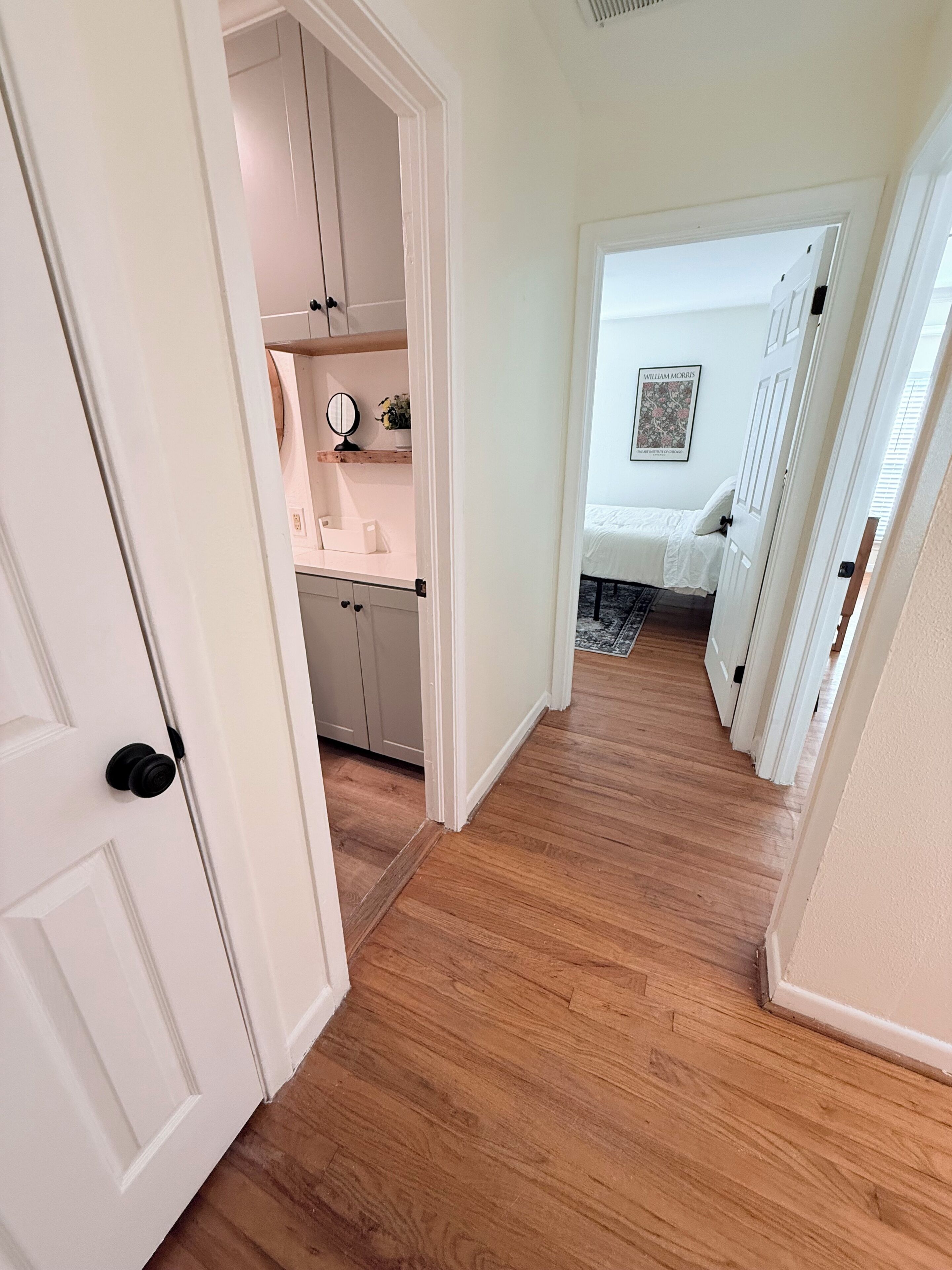 Um corredor com piso de madeira, uma porta branca e um quarto com cama e uma moldura na parede.