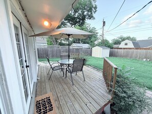 A wooden deck with a table and chairs, a covered umbrella, and a small shed in the background. - Home near DFW Airport & Arlington World Cup. Perfect for groups and families. (Irving)