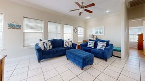 A living room with a sectional sofa, ottoman, ceiling fan, and large windows with blinds. - MR288 Shared Pools, Golf Cart Included, Pet Friendly (Port Aransas)