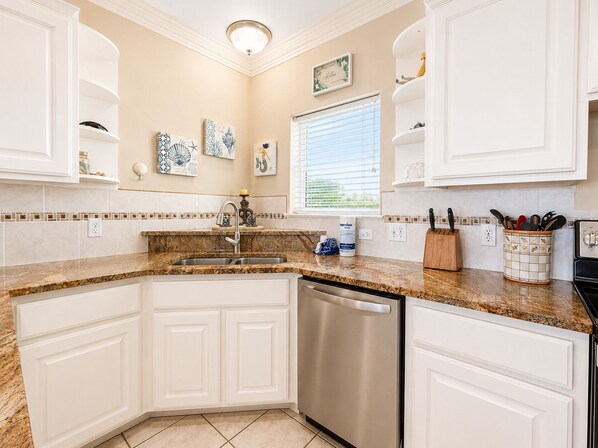 A kitchen with white cabinets, a stainless steel dishwasher, a granite countertop, and a window with blinds. - MR288 Shared Pools, Golf Cart Included, Pet Friendly (Port Aransas)