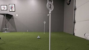 Une pièce dont le sol est recouvert d’un revêtement d’herbe artificielle, avec un poteau cible numéroté et des tableaux encadrés accrochés au mur.