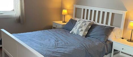 A bedroom with a bed, nightstands, a lamp, a painting, and a window with curtains.