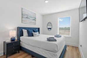 Een slaapkamer met een bed, een nachtkastje, een lamp, een schilderij, een spiegel en een raam met jaloezieën.