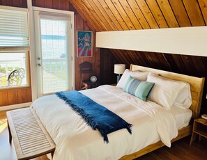 A bedroom with a wooden ceiling, a bed with white bedding and a blue throw, a nightstand with a lamp, and a painting on the wall.