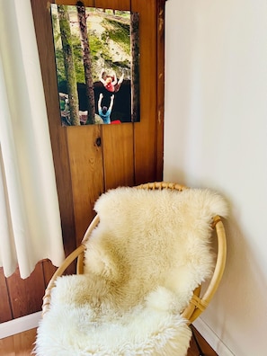 A wicker chair with a fluffy white cushion in a room with wooden walls and a framed photo of a forest scene.