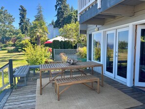 Een houten terras met een tafel en banken, een overdekte patio en uitzicht op een tuin en bomen.