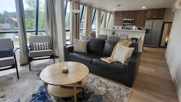 A modern living room with a sofa, coffee table, and chairs, a kitchen in the background, and large windows.
