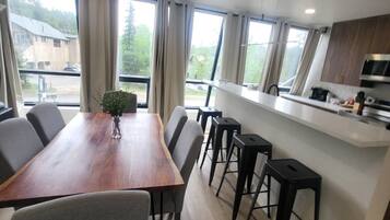 A modern kitchen with a wooden dining table and bar stools, large windows with curtains, and a view of the outside.