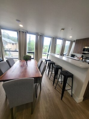 Ett modernt kök med ett matbord och barstolar i trä, stora fönster med gardiner och utsikt över omgivningen.