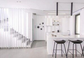 A modern kitchen with white cabinetry, a central island with stools, and a staircase leading to an upper level. - Luxury 2BR Cretan Villa Near Hersonissos Beaches (Hersonissos)