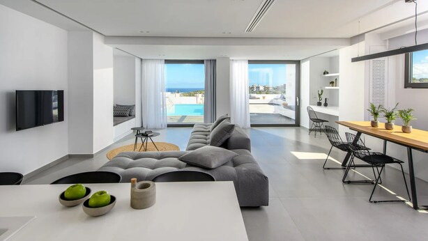 A modern living room with a large gray sofa, a dining area with a wooden table, and a kitchen with a window overlooking a pool. - Luxury 2BR Cretan Villa Near Hersonissos Beaches (Hersonissos)