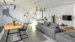 A modern living room with a grey sofa, a wooden dining table with black chairs, a kitchen area, and a staircase. - Luxury 2BR Cretan Villa Near Hersonissos Beaches (Hersonissos)