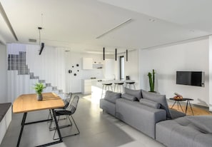 A modern living room with a grey sofa, a wooden dining table with black chairs, a kitchen area, and a staircase.