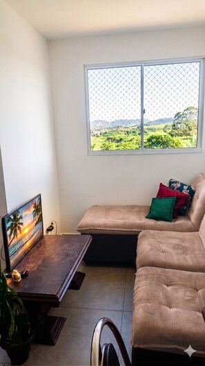 A living room with a sofa, a TV, a window with a view of greenery, and a small table with a plant. - Vrbo Property (Uberaba)
