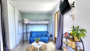 Ein gemütlicher Raum mit Etagenbett, einer Schlafcouch, einem kleinen Tisch mit Pflanze, einem Bücherregal und einem Fenster mit Vorhängen.