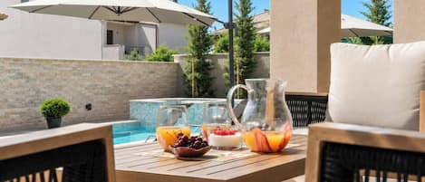 A poolside area with a wooden table, two chairs, a pitcher of fruit, and a glass of orange juice.