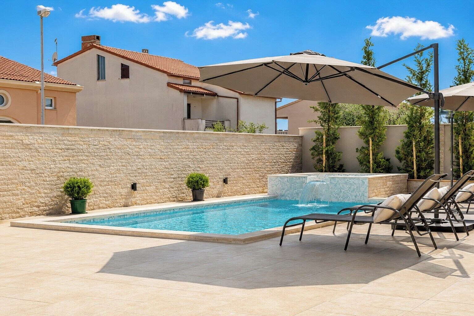 A swimming pool with lounge chairs and an umbrella, surrounded by stone walls and potted plants.