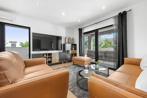 Un salon moderne avec des canapés en cuir, un téléviseur à écran plat, une table basse et une porte-fenêtre coulissante donnant sur l’extérieur.