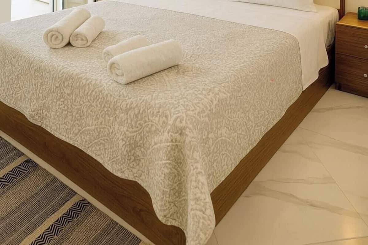 Una cama bien hecha con una colcha y almohadas estampadas, un cabecero de madera y vista al mar a través de una puerta abierta.